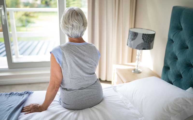 An older woman with gray hair sitting on a bed. She is seen from the back, looking out her window.